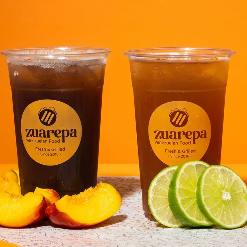Two iced beverages labeled Zuarepa Venezuelan Food, surrounded by sliced peaches and limes against a bright orange background.