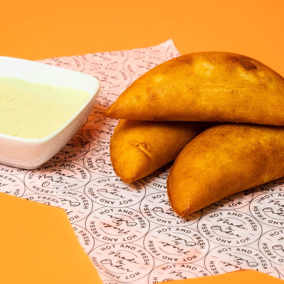 Three golden fried pastries served with creamy green dipping sauce on branded paper against an orange background.