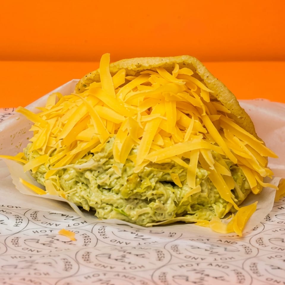 Crunchy taco filled with guacamole and topped with abundant shredded cheddar cheese on branded paper.