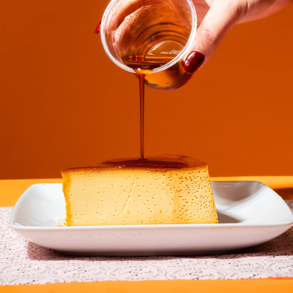 Hand pours amber syrup over a golden, porous cake slice on a white plate against an orange background.
