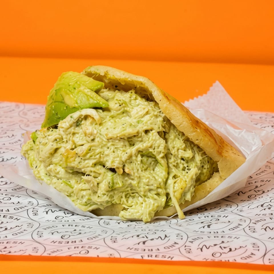 Avocado chicken salad sandwich on toasted bread, wrapped in branded paper against a bright orange background.