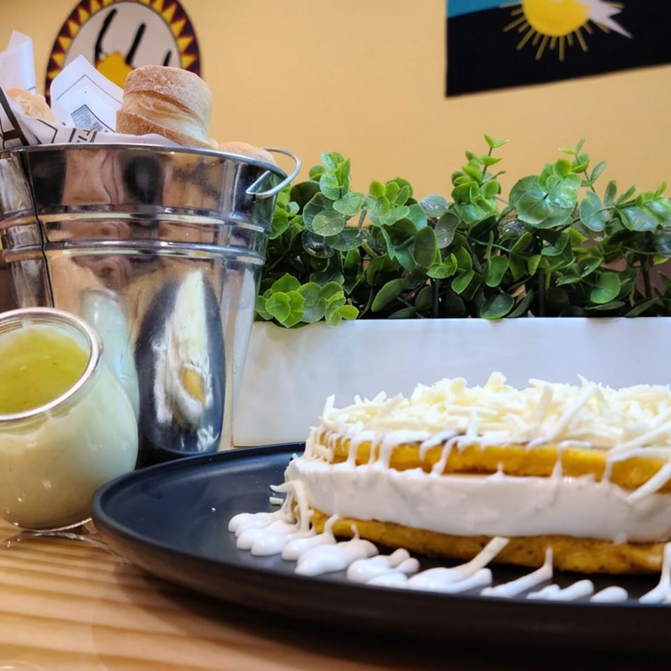 Layered golden cornmeal dish topped with shredded white cheese on a black plate, beside sauce, bread, and greenery in a restaurant.