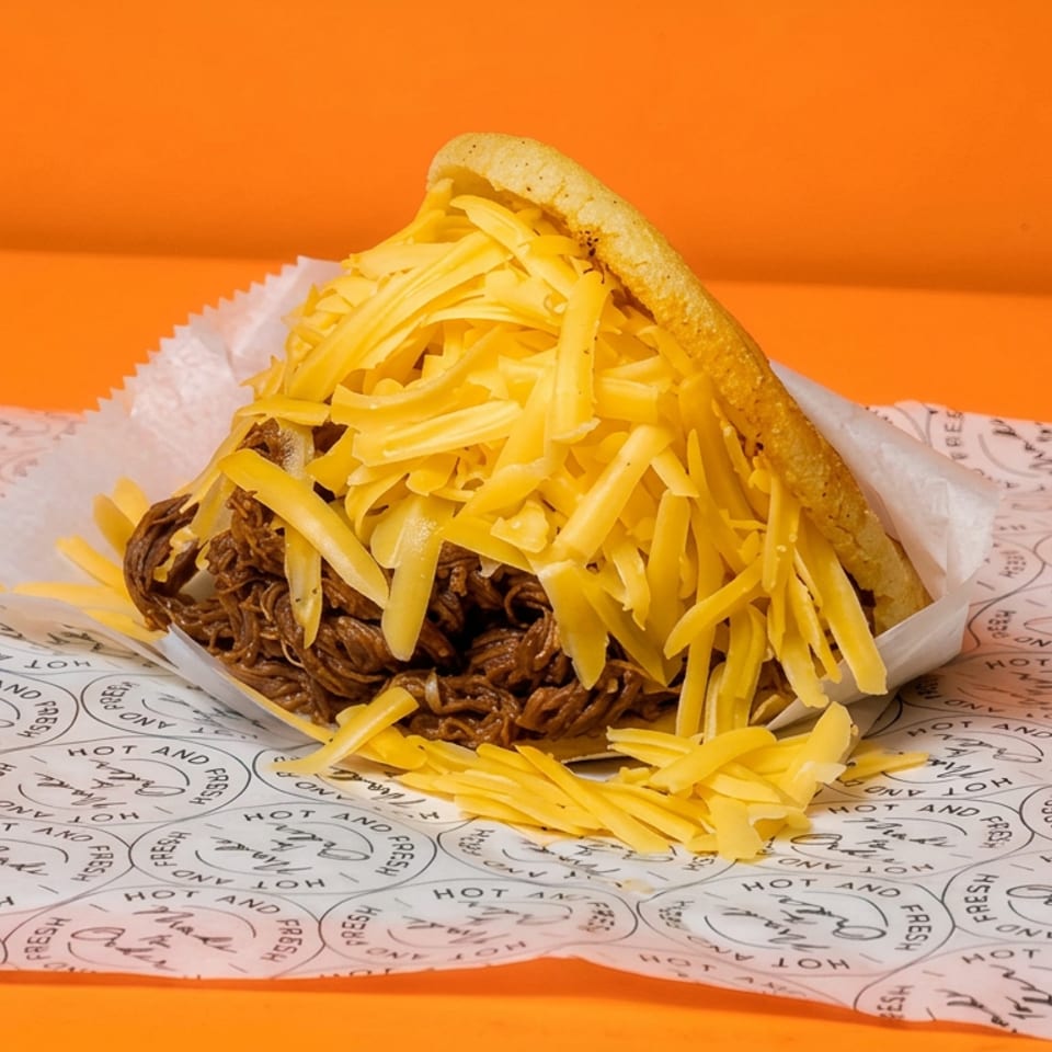 Open pita sandwich filled with shredded meat and abundant grated cheese on branded paper against an orange background.