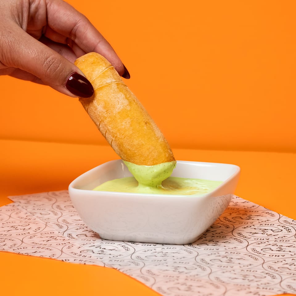 Hand dips a golden fried snack into creamy green sauce in a white bowl against an orange background.