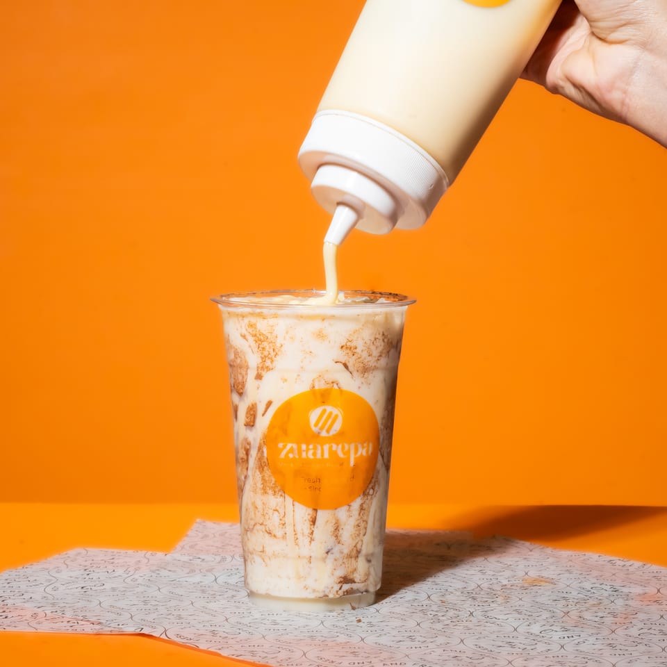 Hand pours creamy sauce from a squeeze bottle into a branded dessert cup against a bright orange background.