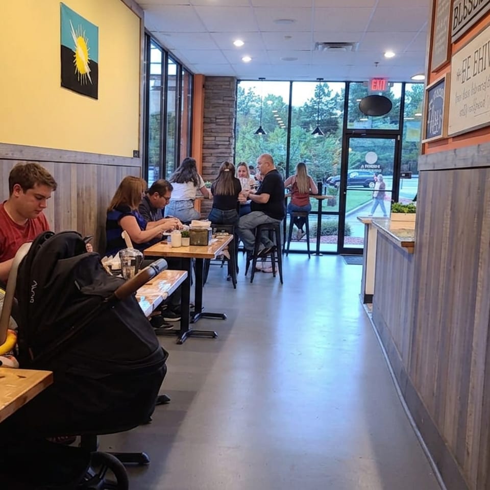 Casual restaurant interior with diners at wooden tables, a stroller, colorful wall artwork, and large windows overlooking trees.