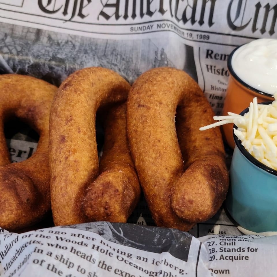 Three golden fried dough rings served with creamy sauce and shredded cheese in colorful cups on newspaper-lined basket.