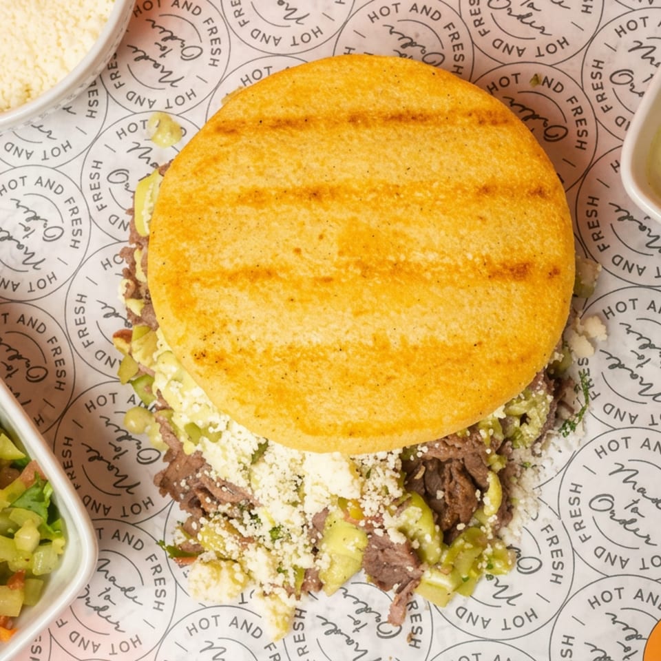 Overhead view of a golden arepa stuffed with shredded beef, avocado, and cheese, served with salsa, white sauce, and grated cheese.